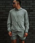 Person wearing a light gray jacket and shorts standing against a dark brick wall.