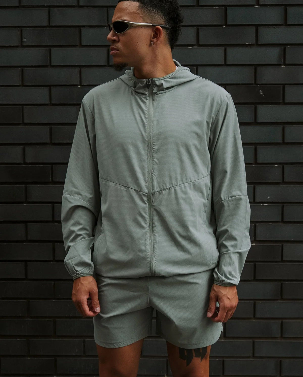 Person wearing a light gray jacket and shorts standing against a dark brick wall.