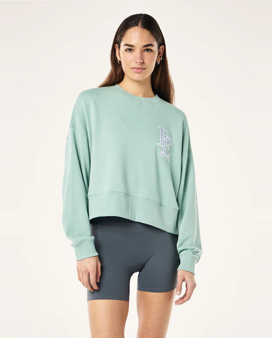 Woman wearing a light green sweatshirt with a logo and dark gray shorts on a white background