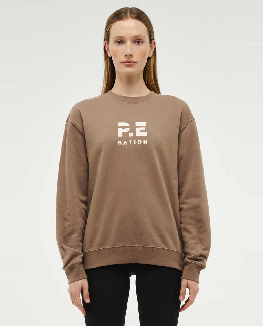 A woman wearing a beige crew neck sweatshirt with 'P.E NATION' branding on the front.