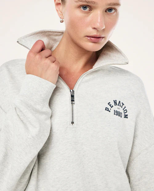 Person wearing a light gray jacket with 'P.E. Nation 1980' branding on a white background