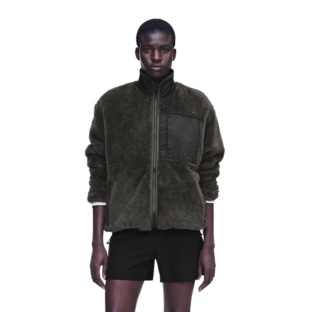 A model wearing a dark olive green sherpa zip-up jacket with a black chest pocket and black shorts, photographed against a solid white background.