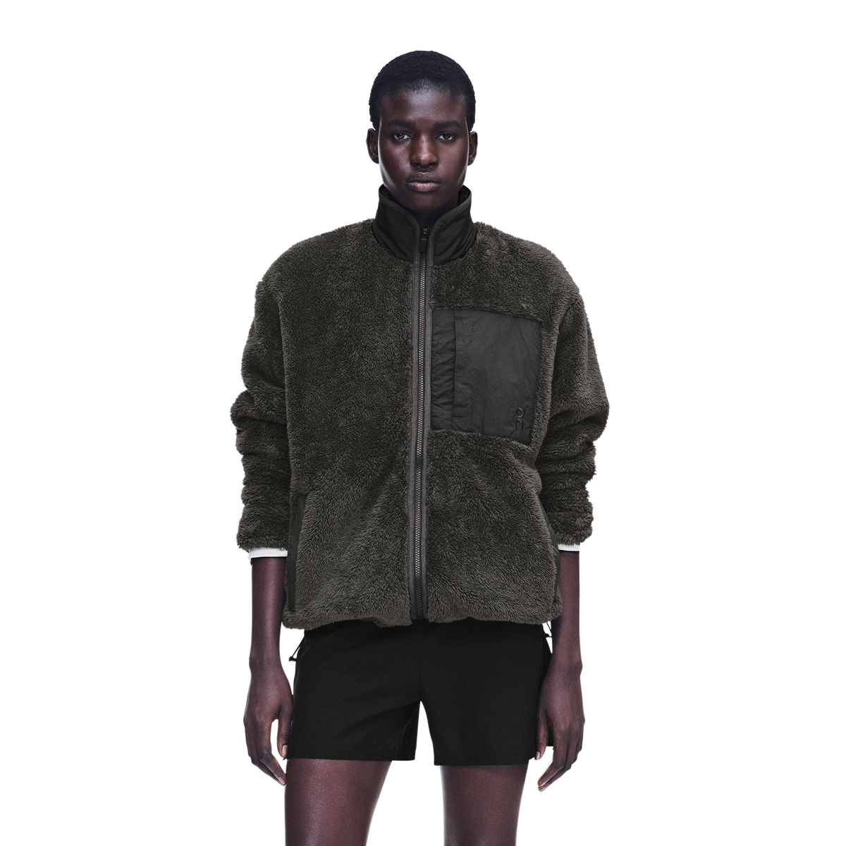 A model wearing a dark olive green sherpa zip-up jacket with a black chest pocket and black shorts, photographed against a solid white background.