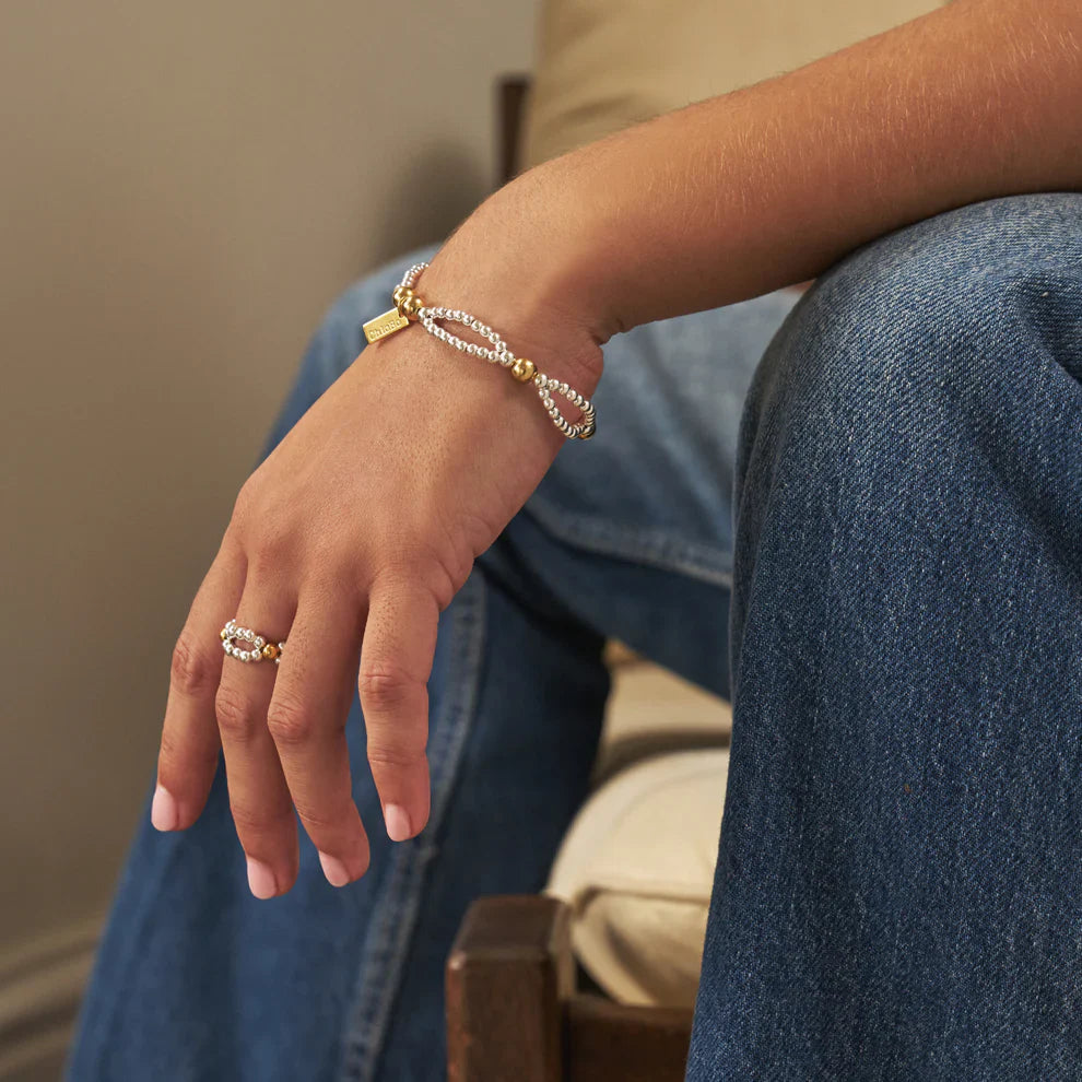 A person wearing blue jeans sits in a chair and shows off gold and silver beaded jewelry.
