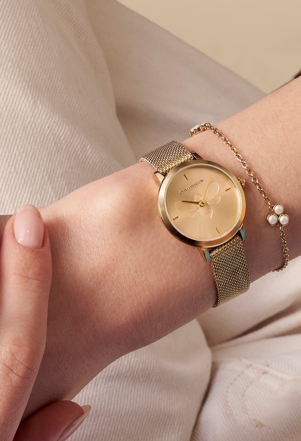 Wrist view of Olivia Burton gold-plated mesh watch with bee detail, styled with a delicate gold bracelet.