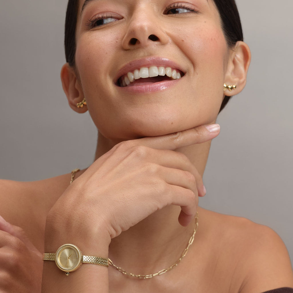 A smiling woman with her hand to her chin, wearing gold jewelry and a watch.
