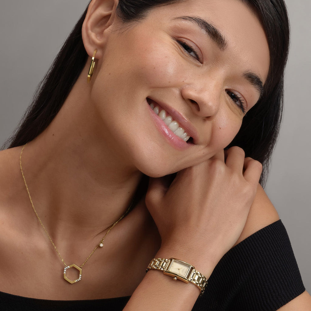 Smiling woman wearing gold earrings, necklace, and watch. 
