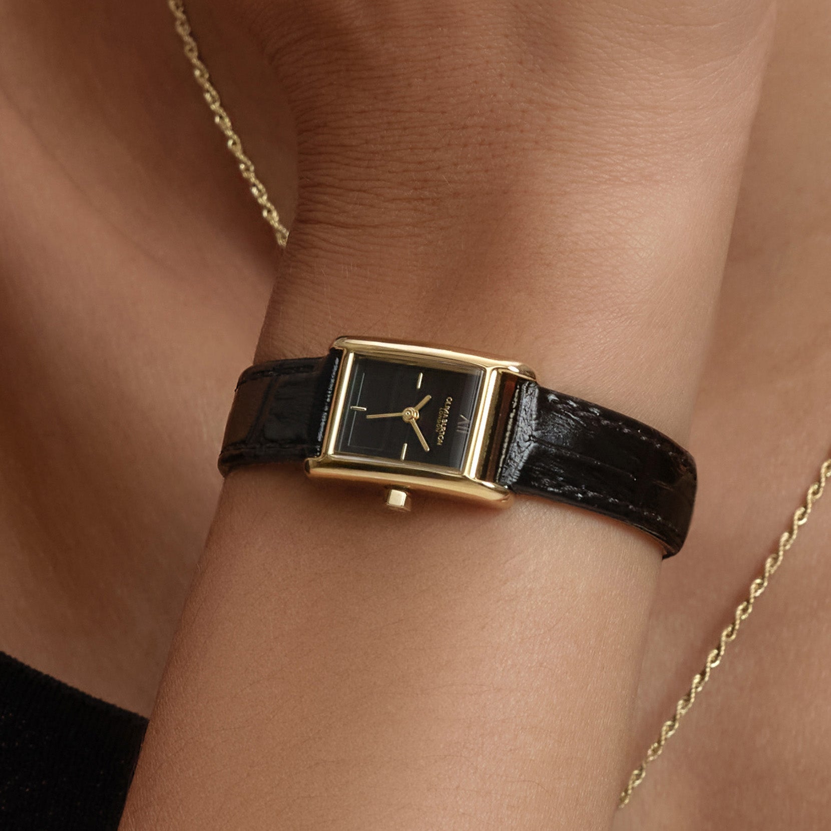 Close-up of a woman's wrist wearing a gold-toned rectangular watch with a black leather strap, set against skin and gold chain.
