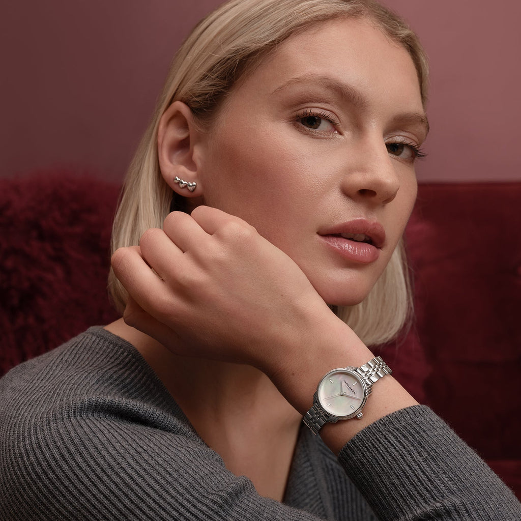 Woman wearing a silver wristwatch and silver heart-shaped earrings, gray sweater, reddish background.
