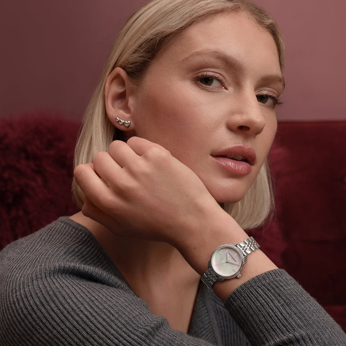 Woman wearing a silver wristwatch and silver heart-shaped earrings, gray sweater, reddish background.
