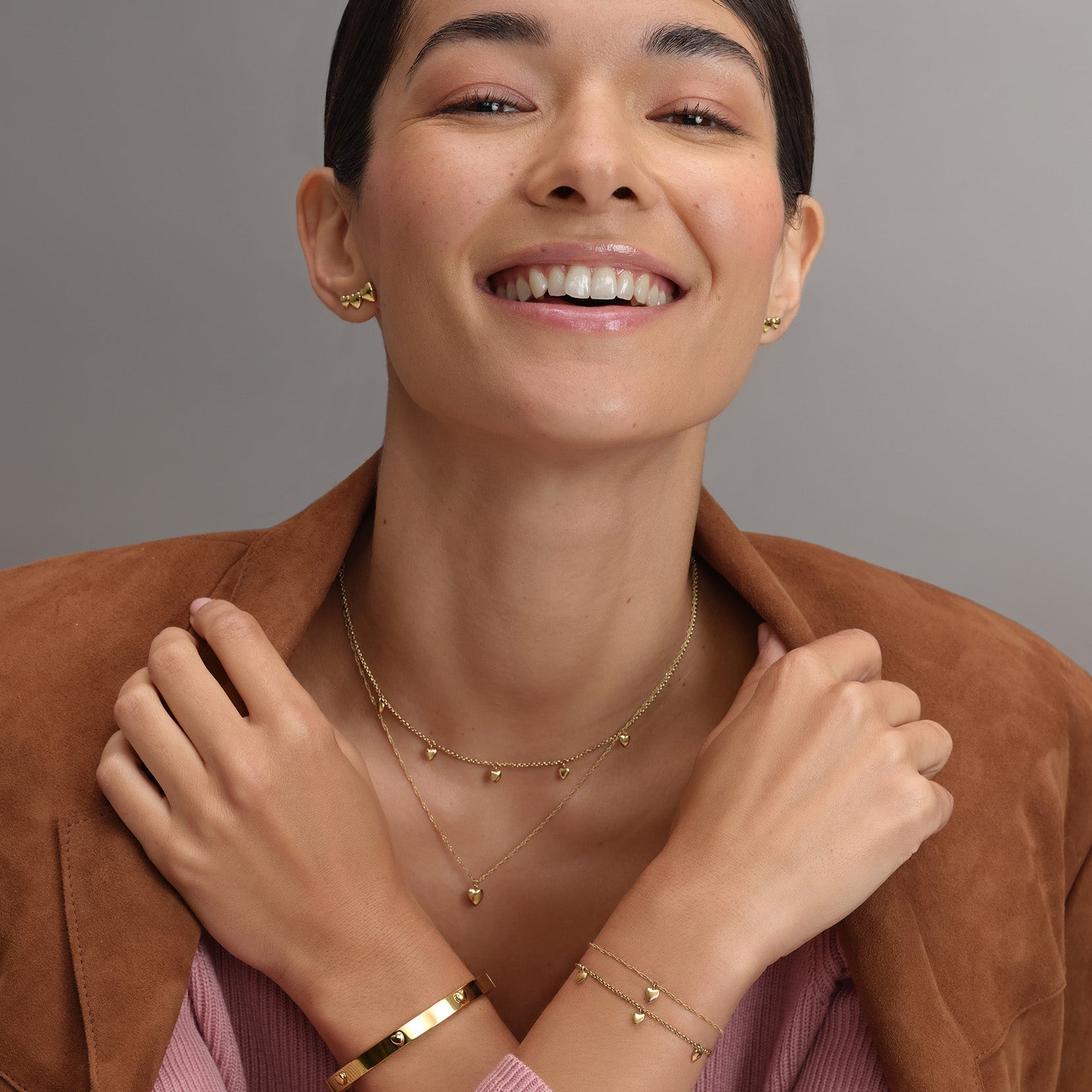 A woman smiling while wearing gold jewelry and a brown jacket.