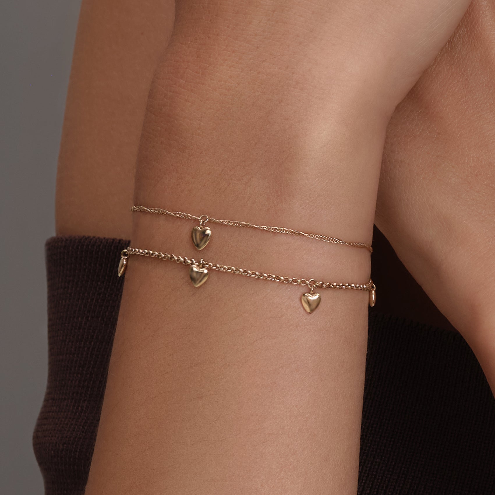 Close-up of two gold heart charm armlets on a tan-skinned arm with a brown shirt sleeve.
