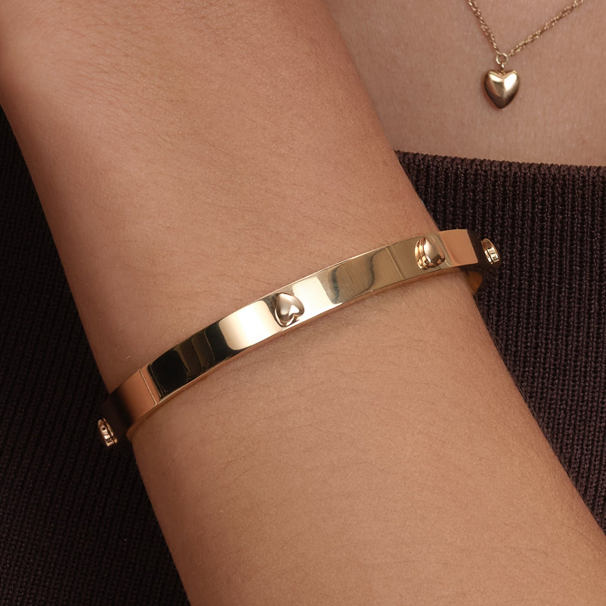 A gold bracelet with heart details worn on a wrist, paired with a gold heart pendant.
