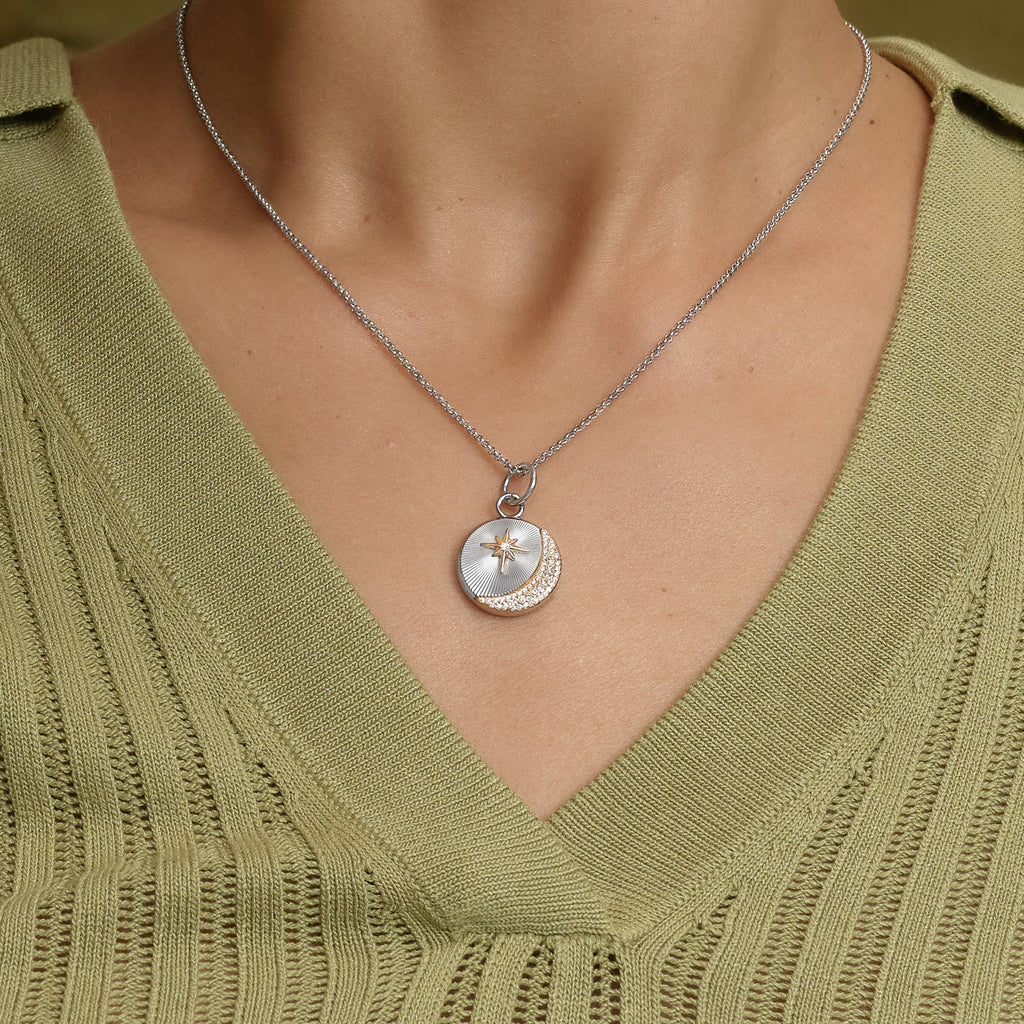 Woman wearing a silver necklace with a circular pendant that features a star and crescent moon design, over a light green knitted top with a V-neck.