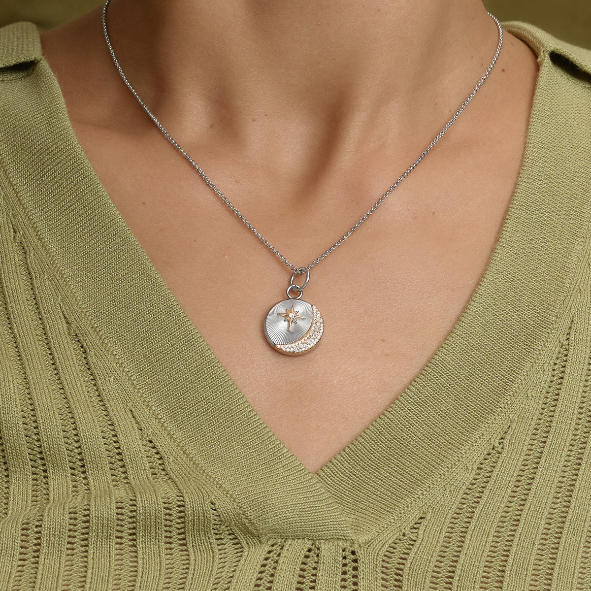 Woman wearing a silver necklace with a circular pendant that features a star and crescent moon design, over a light green knitted top with a V-neck.