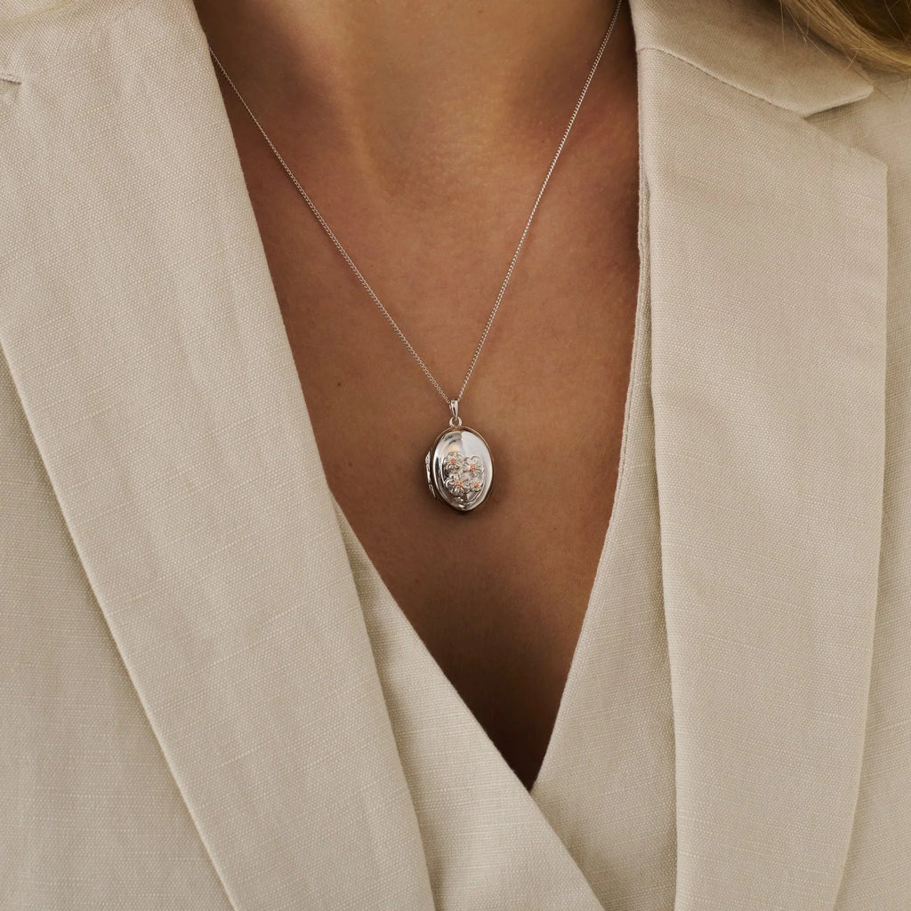 Close-up of a woman wearing a silver locket necklace with floral embellishments against a textured, beige blazer.