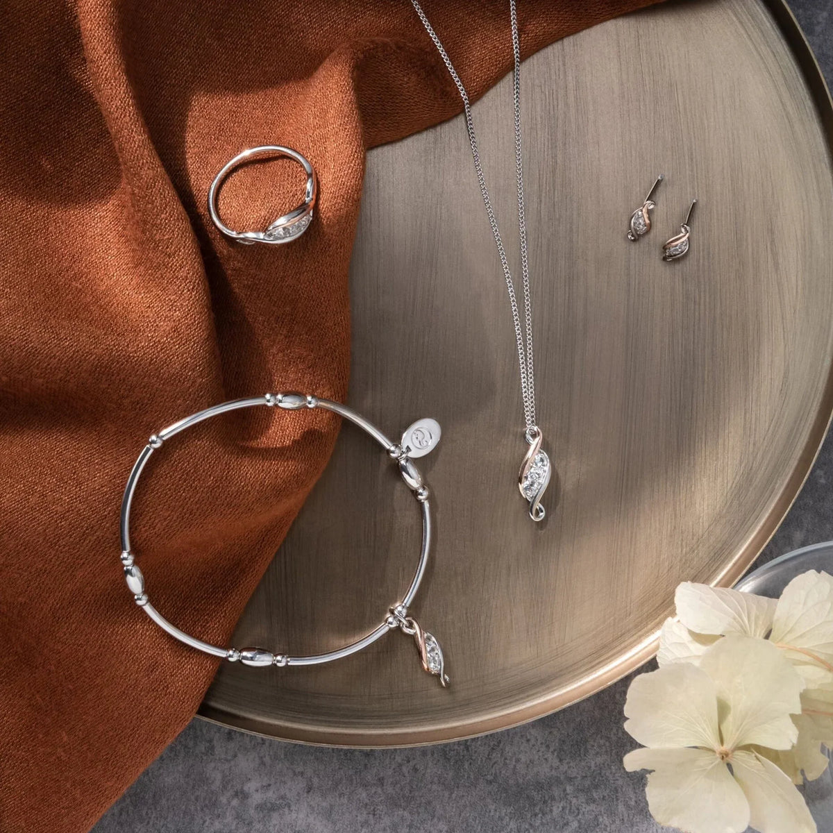 A collection of jewelry, including a ring, bracelet, necklace, and earrings, is displayed on a round metallic tray alongside a brown cloth and white flowers.