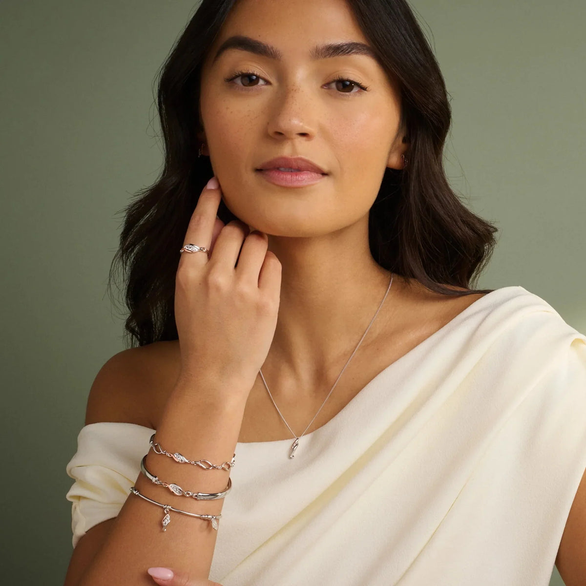 A woman modeling jewelry against a green background, wearing a cream-colored dress with one shoulder exposed.
