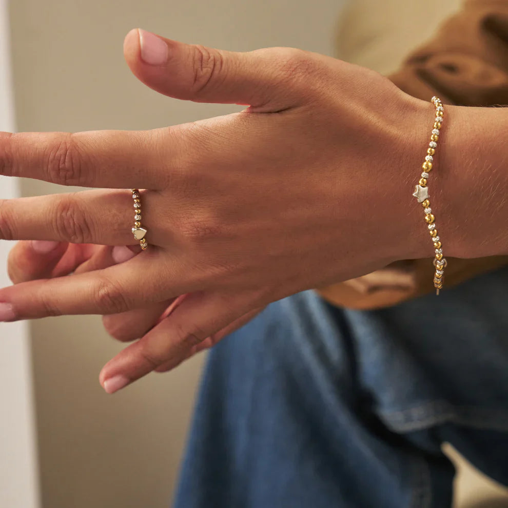 A hand wearing a gold beaded ring and bracelet with star charms.
