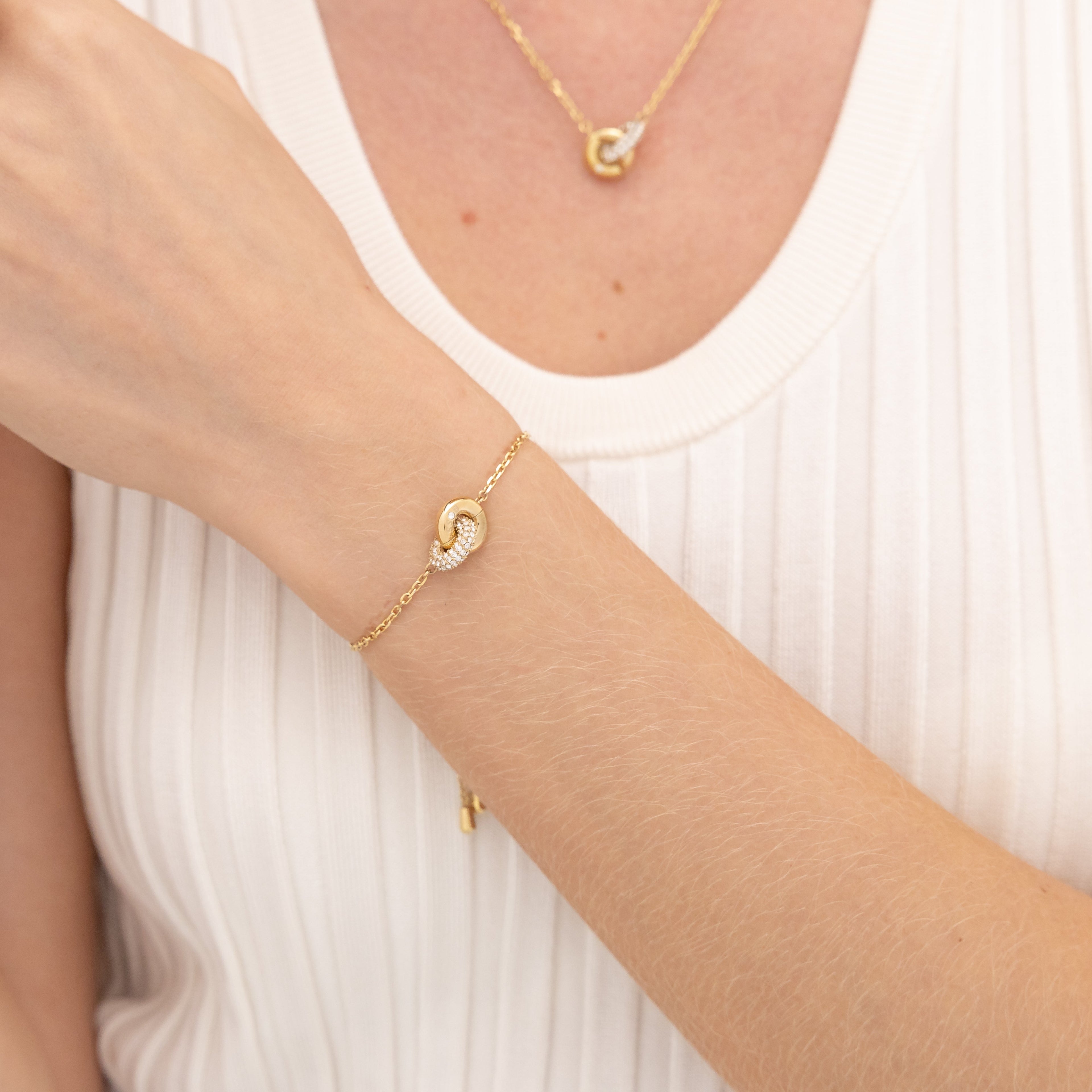 A fair-skinned person wearing a gold bracelet with a linked circle design on their left wrist, paired with a matching necklace, poses against a white ribbed top.
