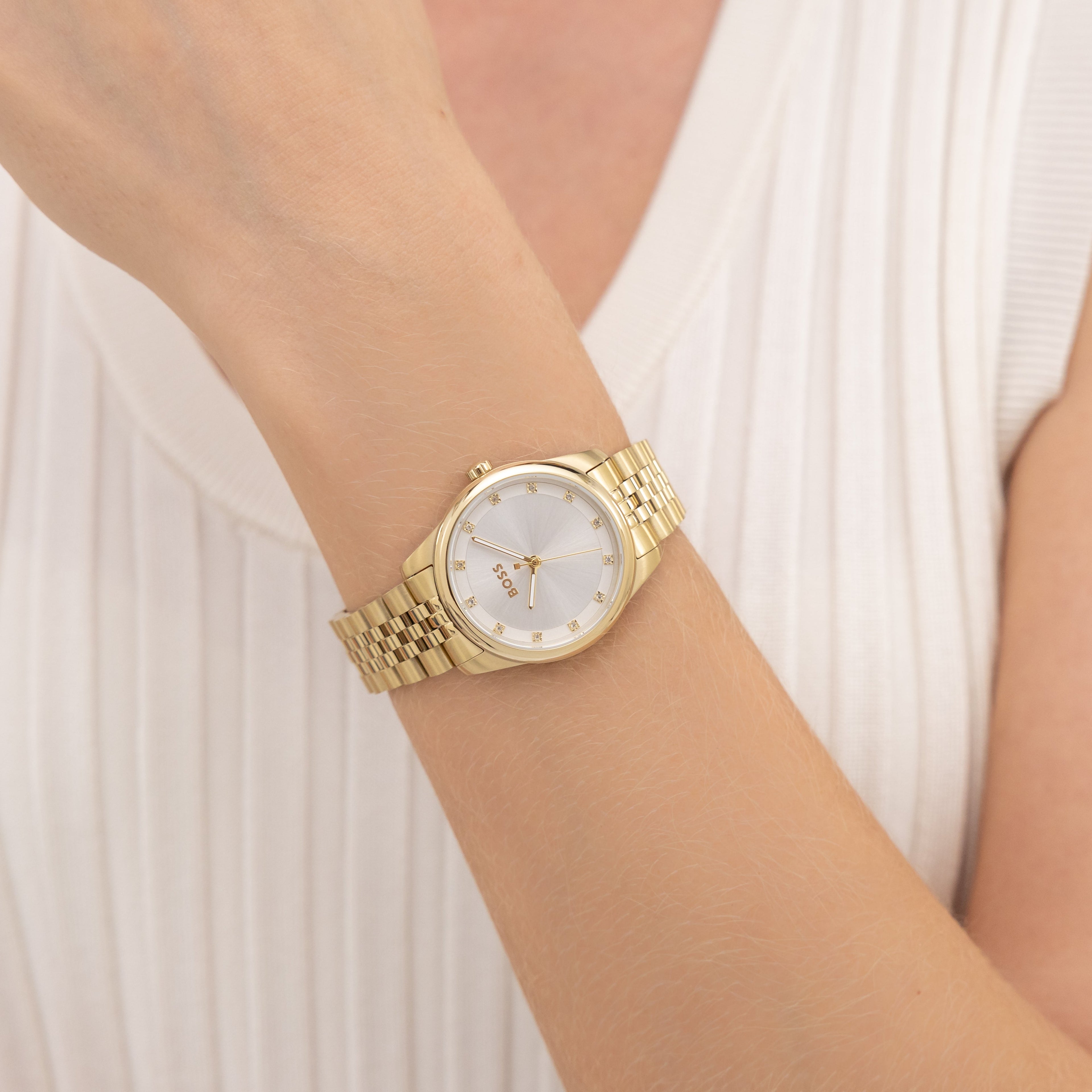 Close-up of a woman's arm wearing a gold watch with a silver face and the brand "BOSS" displayed on the face, against a white, pleated top.
