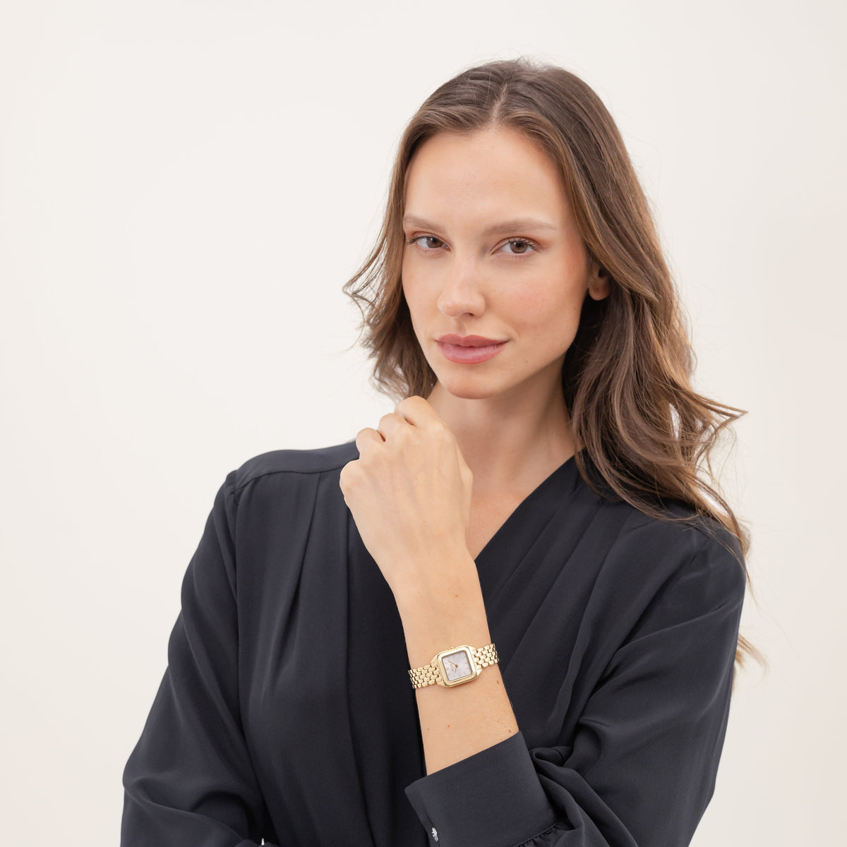 Woman wearing a gold wristwatch, posing in a dark blouse against a neutral background.
