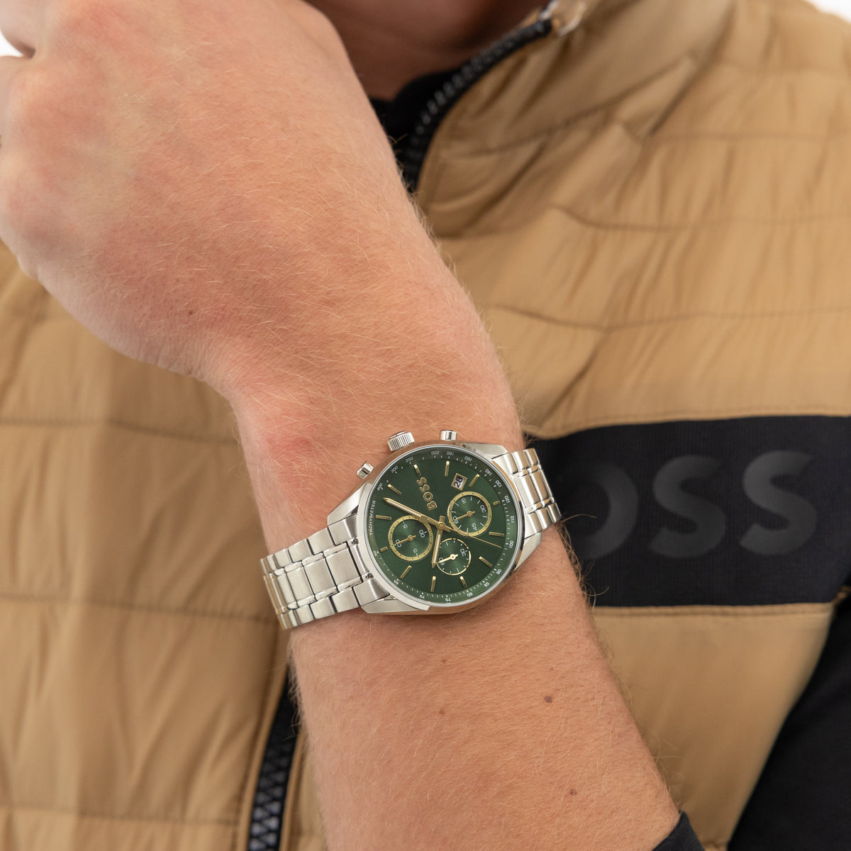A man's arm wearing a silver Boss watch with a green face, set against a beige vest and black shirt.