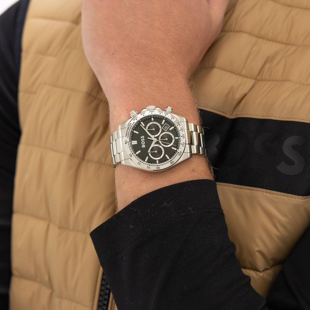 A close-up shot of a man's wrist wearing a silver Hugo Boss wristwatch. The man is wearing a tan vest with a black undershirt.