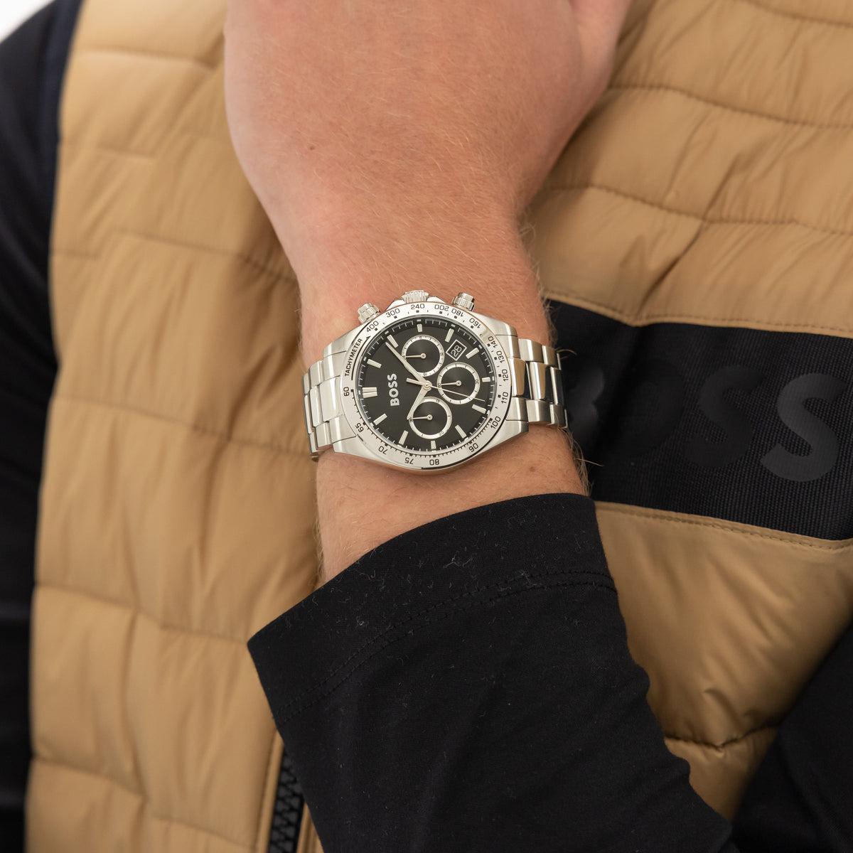 A close-up shot of a man's wrist wearing a silver Hugo Boss wristwatch. The man is wearing a tan vest with a black undershirt.