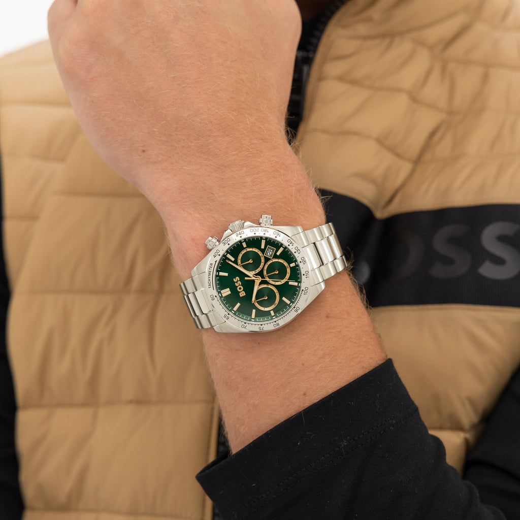 A close-up shot of a man's wrist wearing a silver BOSS wristwatch with a green dial and gold subdials, set against a tan and black BOSS puffer vest.
