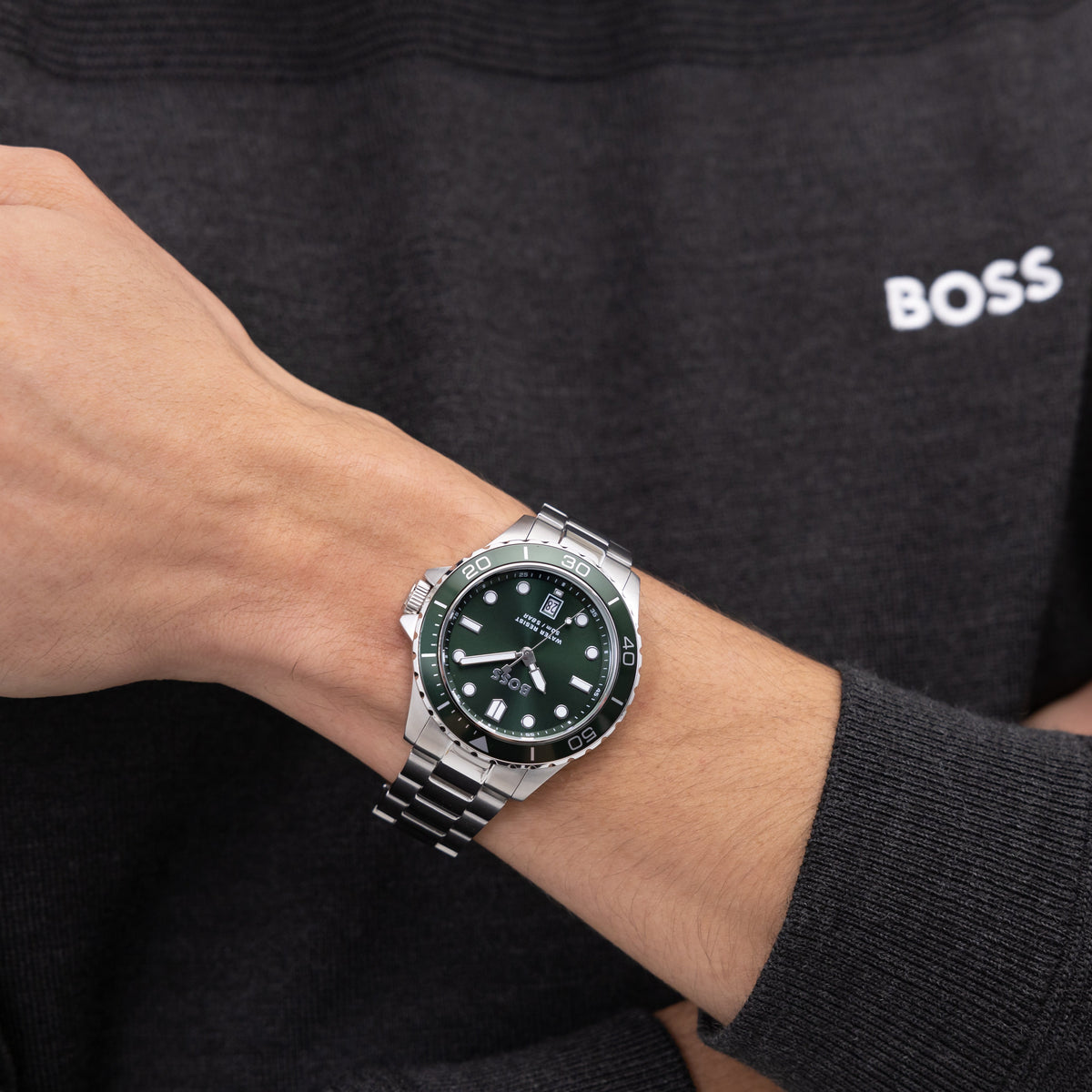 Close-up of a wrist wearing a silver Boss watch with a green face and a dark gray sweater.
