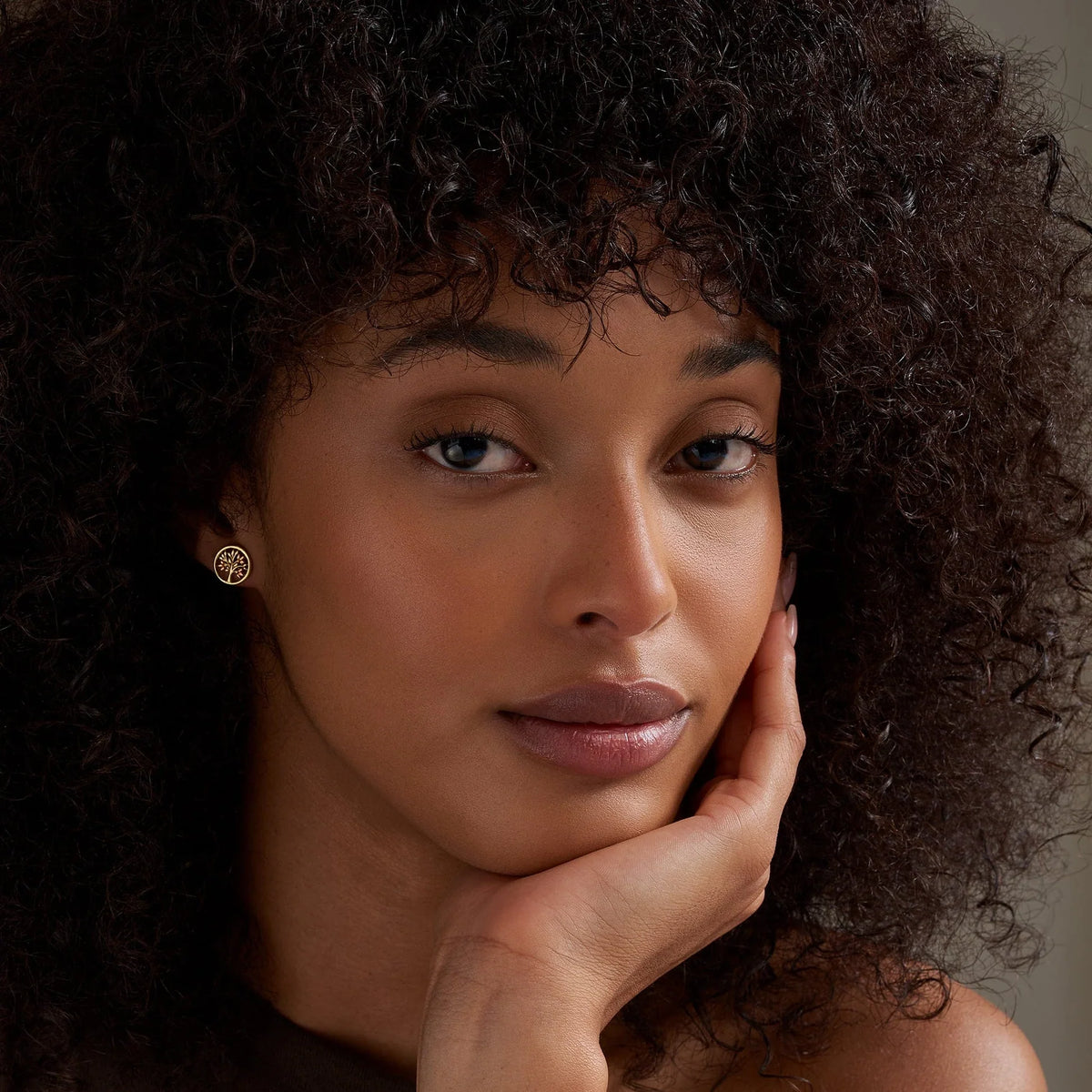 Woman with curly hair poses with hand on chin, wearing gold tree-of-life earrings.