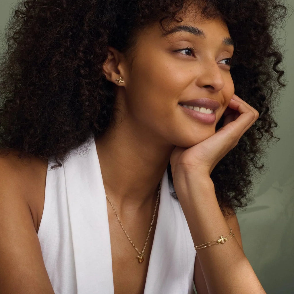 Woman with dark curly hair wearing bee earrings, necklace, and bracelet, resting her head on her hand. She is wearing a white top.