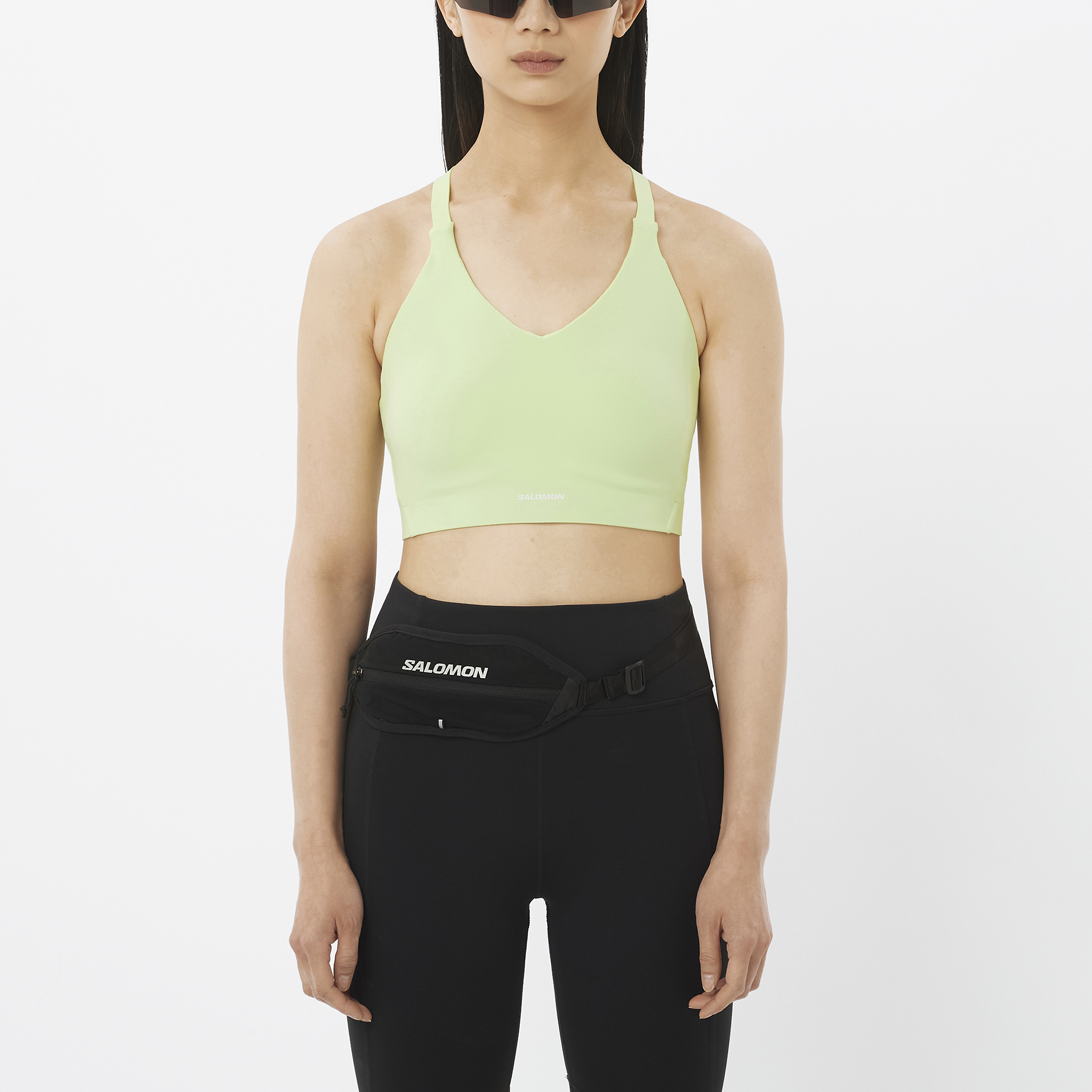 Woman wearing a light green sports bra and black leggings with a Salomon waist bag on a white background