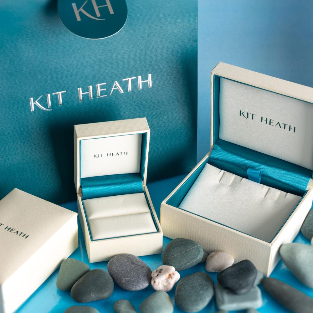 Product shot of Kit Heath branded jewelry boxes and bag, surrounded by gray and white stones on a blue background.