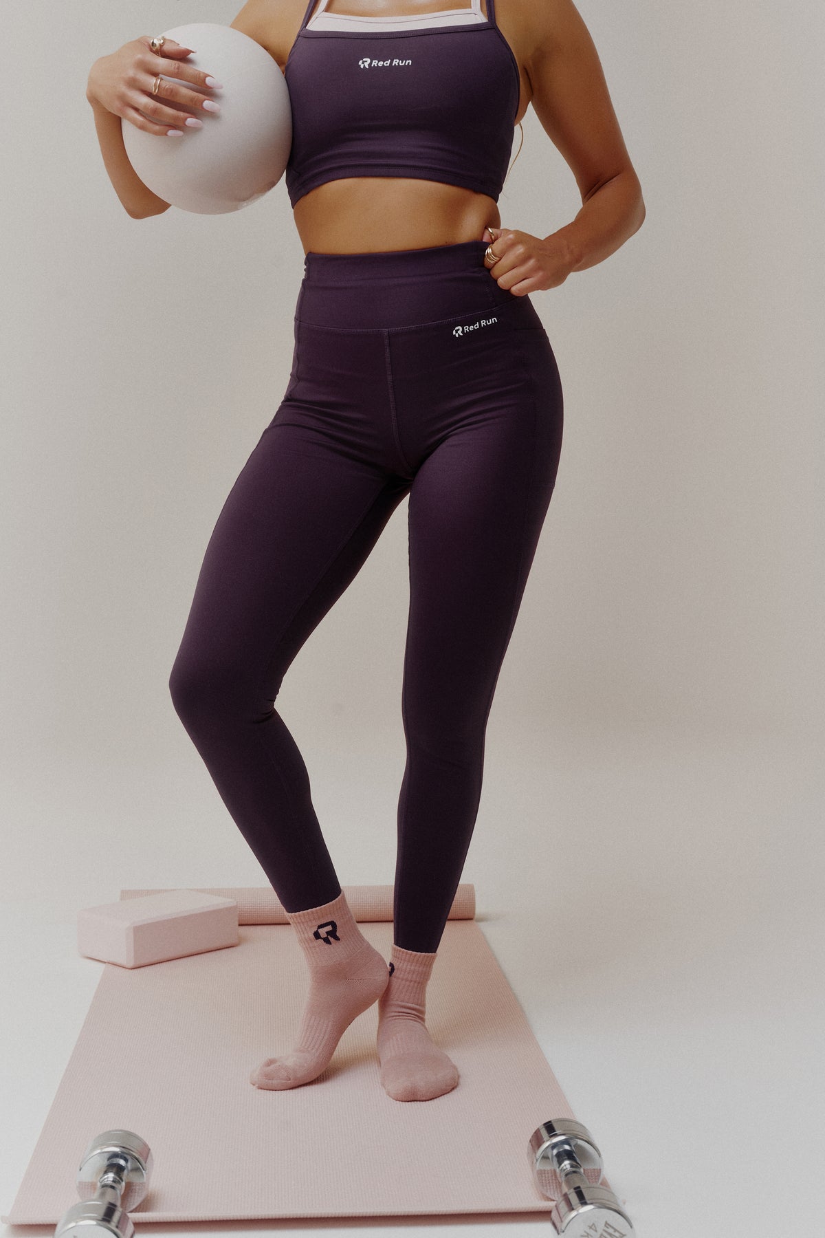 A woman modeling a matching dark purple activewear set consisting of a sports bra and high-waisted leggings, standing on a pink yoga mat with dumbbells and yoga blocks in a studio setting.