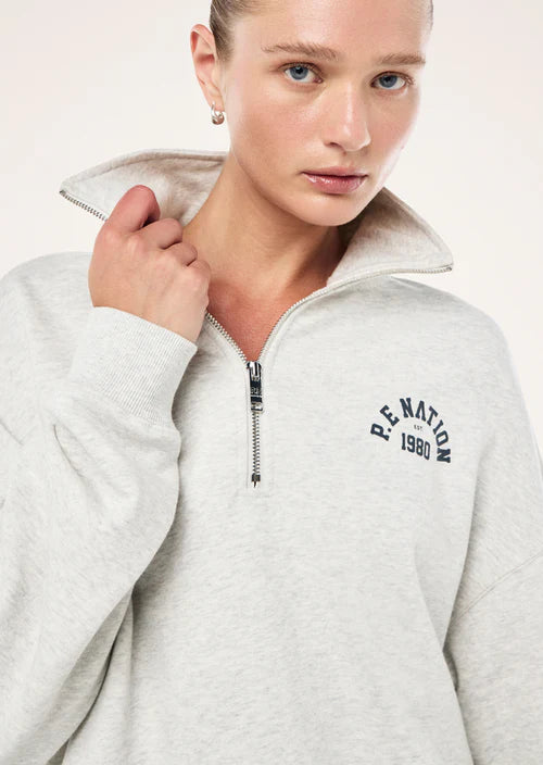 Person wearing a light gray jacket with 'P.E. Nation 1980' branding on a white background