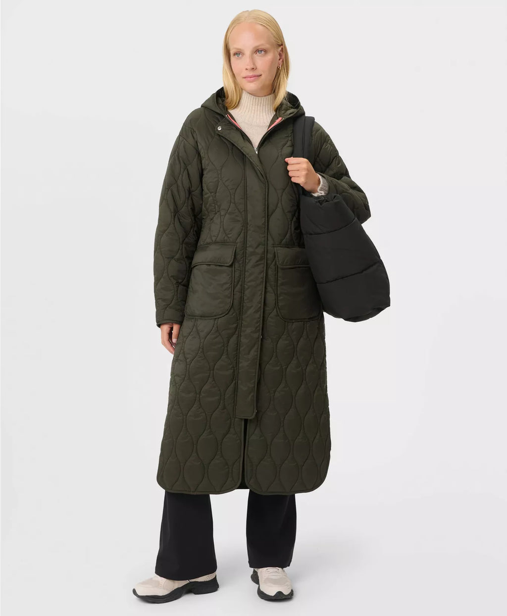 Person wearing a long quilted green coat holding a black bag on a white background