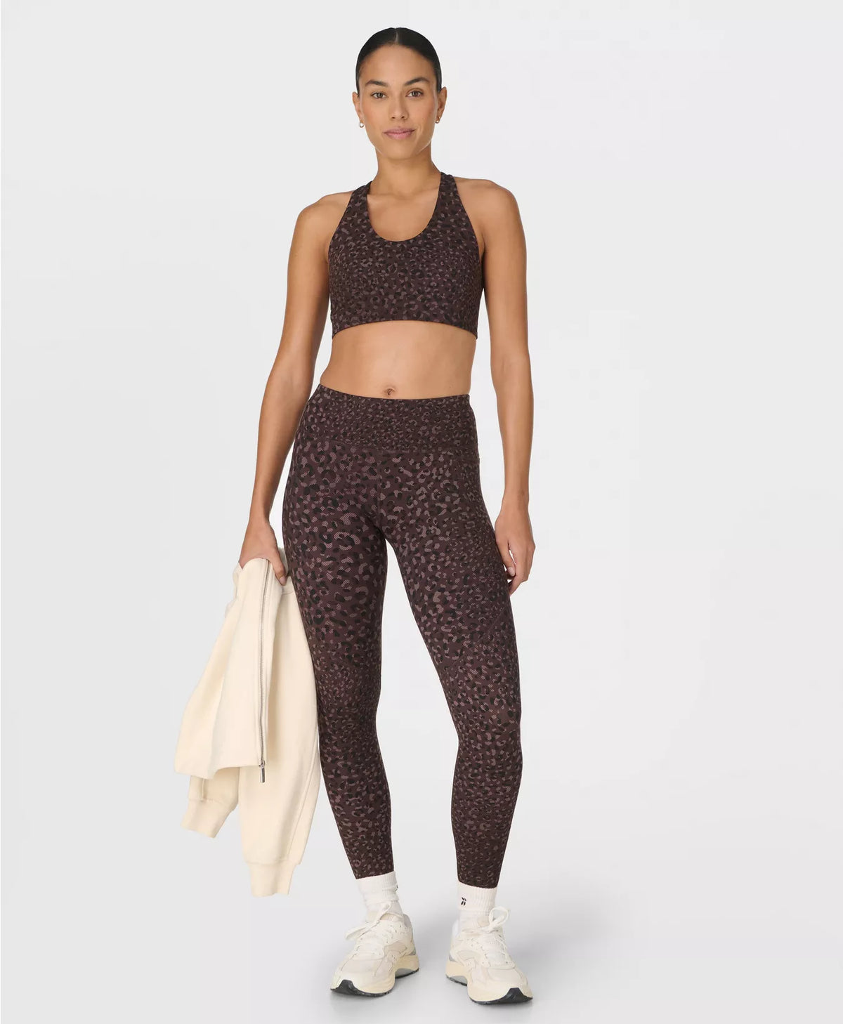 Woman wearing a patterned sports bra and leggings holding a beige jacket on a white background