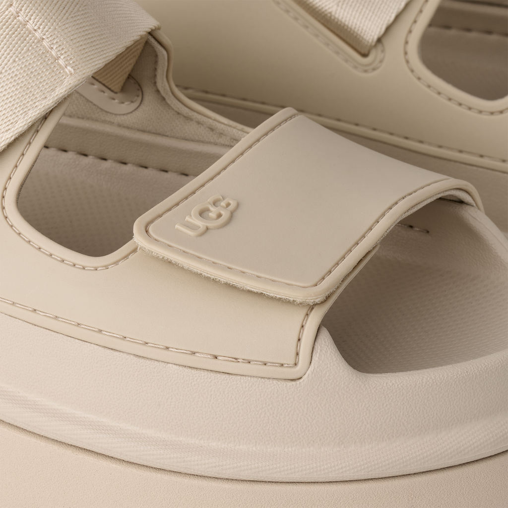Close-up of a beige sandal with a strap and UGG logo.