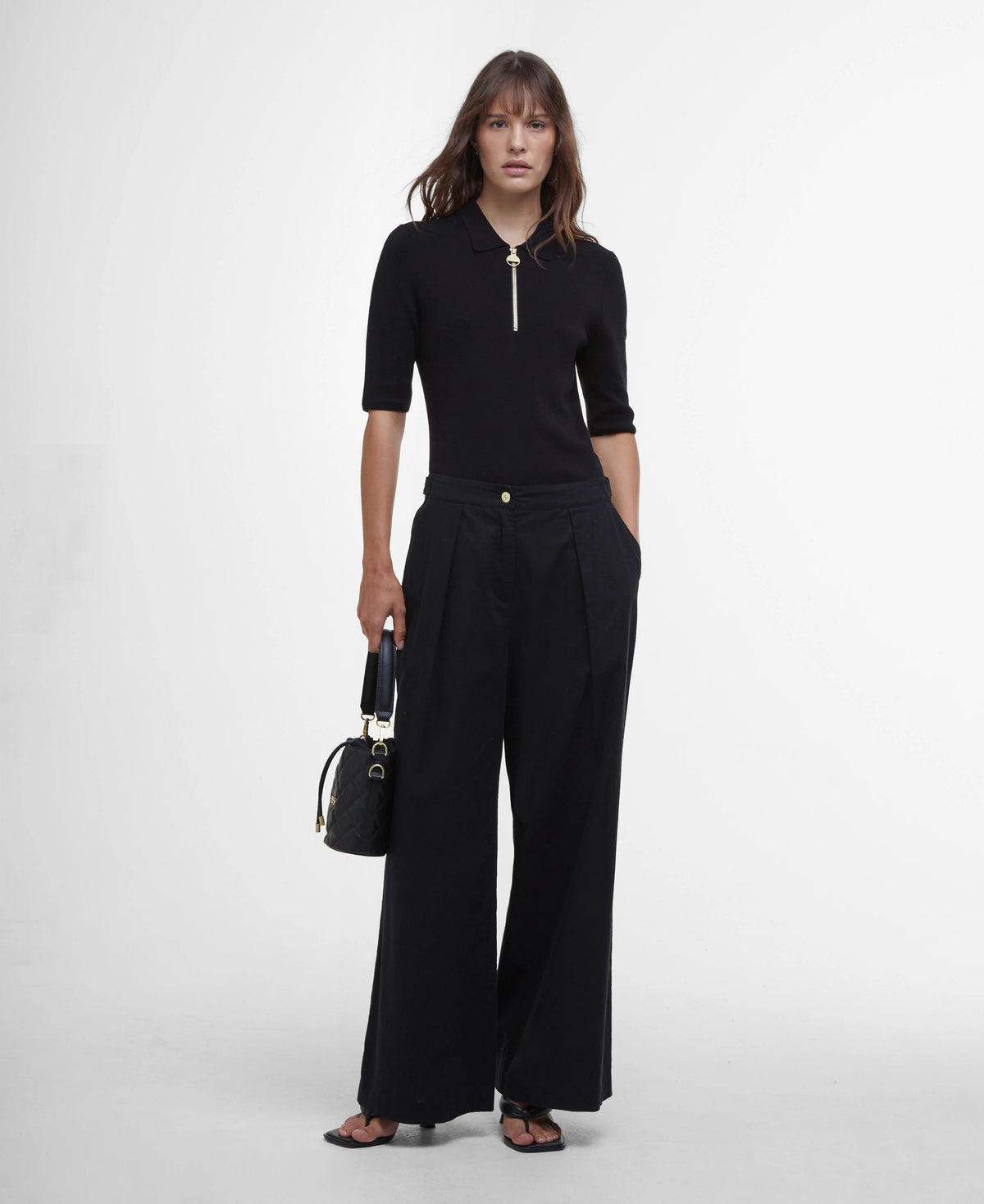 A woman modeling Barbour Black Riley Trousers, which are black in color, made from a cotton and linen blend, featuring an adjustable waist with branded studs, side and back pockets, and a silicone ID badge.