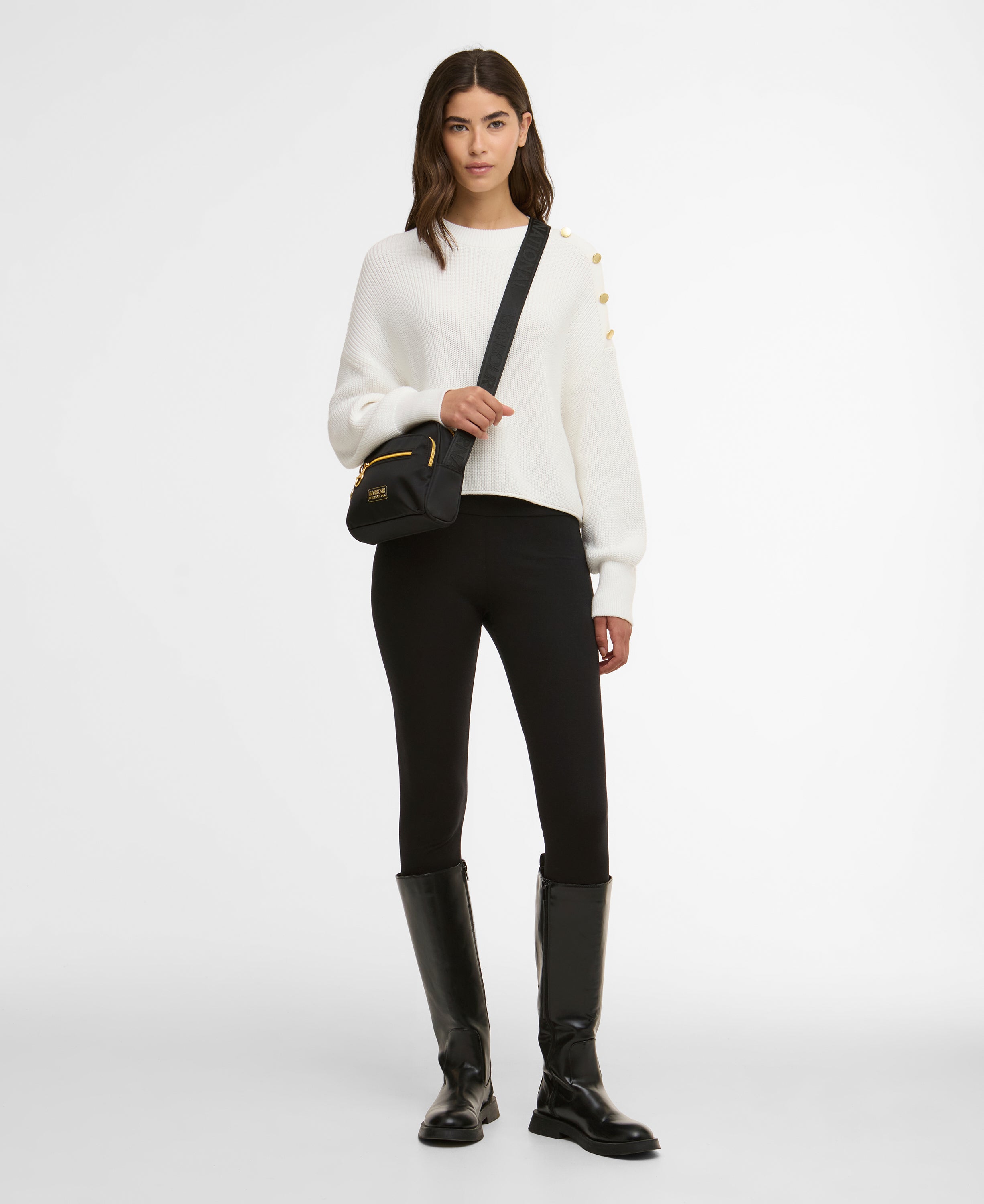 Woman wearing a white jumper, black pants, and black boots holding a black bag on a white background
