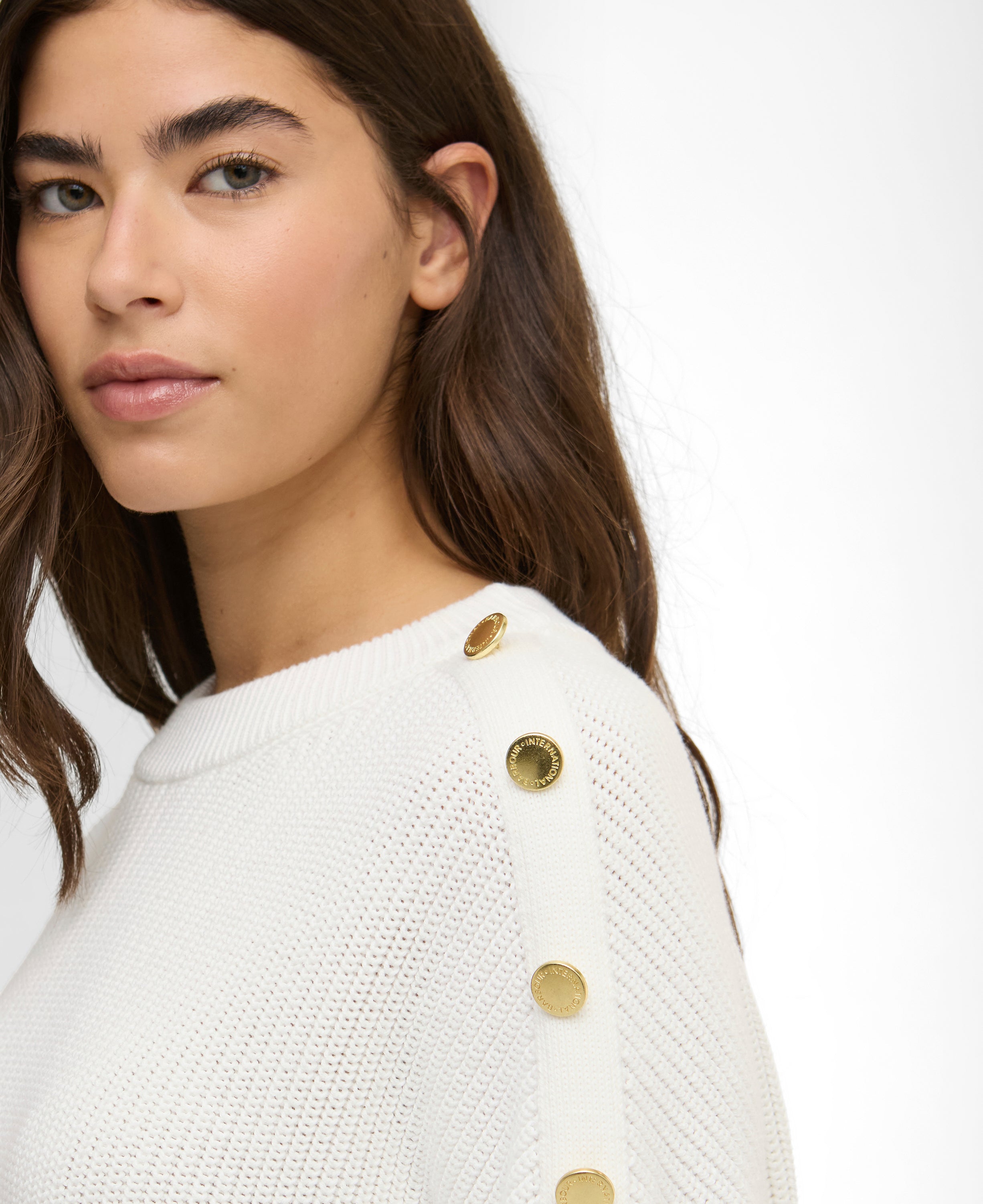 Woman wearing a white sweater with gold buttons on a plain background