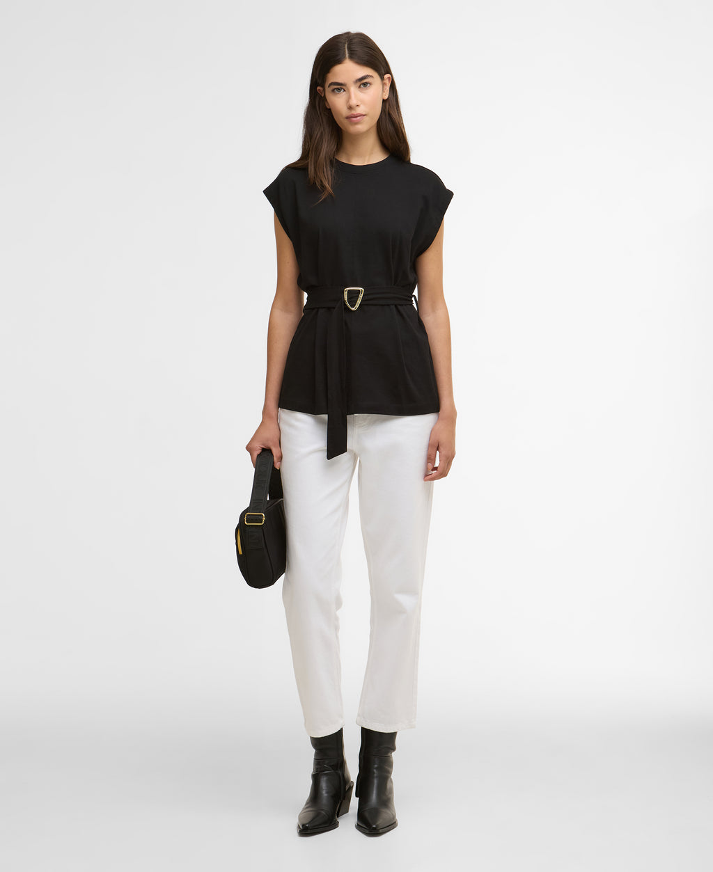 Woman wearing a black top and white pants with a belt, holding a black handbag on a white background