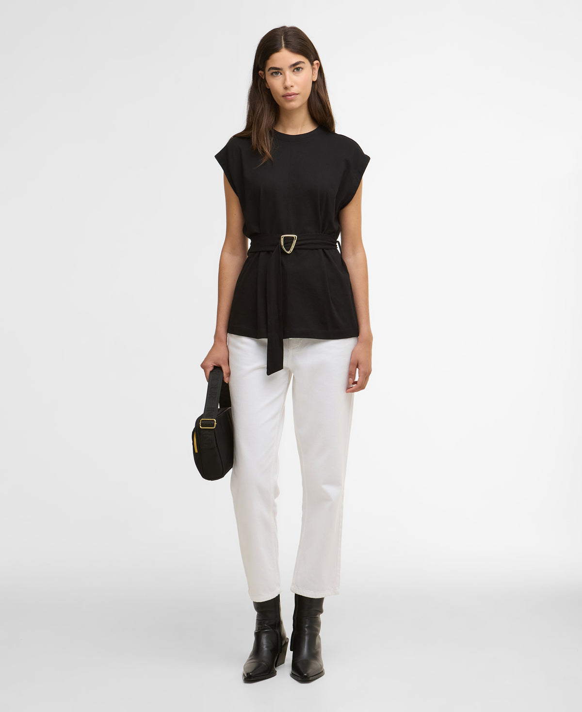 Woman wearing a black top and white pants with a belt, holding a black handbag on a white background