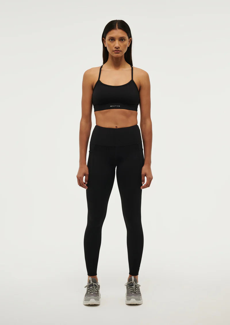 A woman standing, wearing black high-waisted leggings with a drawcord and a sports bra. She is also wearing sneakers.