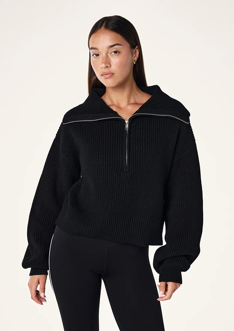 A woman wearing a black zip neck knit sweater with long sleeves, from the brand P.E Nation.