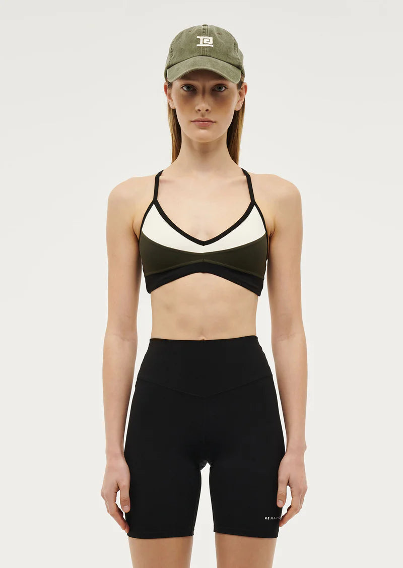 A woman wearing a khaki sports bra with a cross back design and thin adjustable straps. The bra features contrast panels and a V neckline.