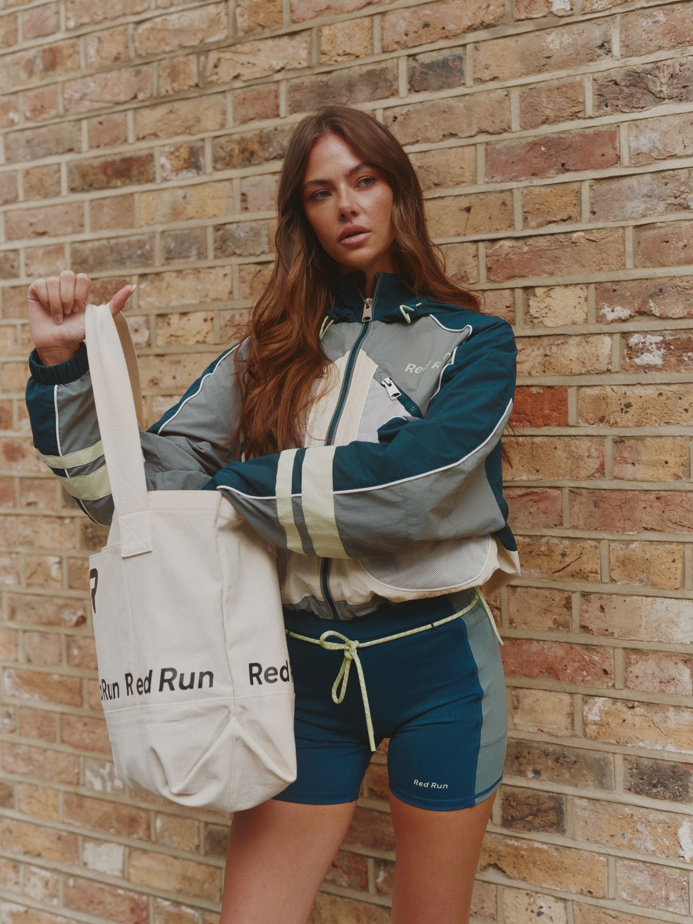 Woman holding a tote bag against a brick wall wearing a sports outfit.