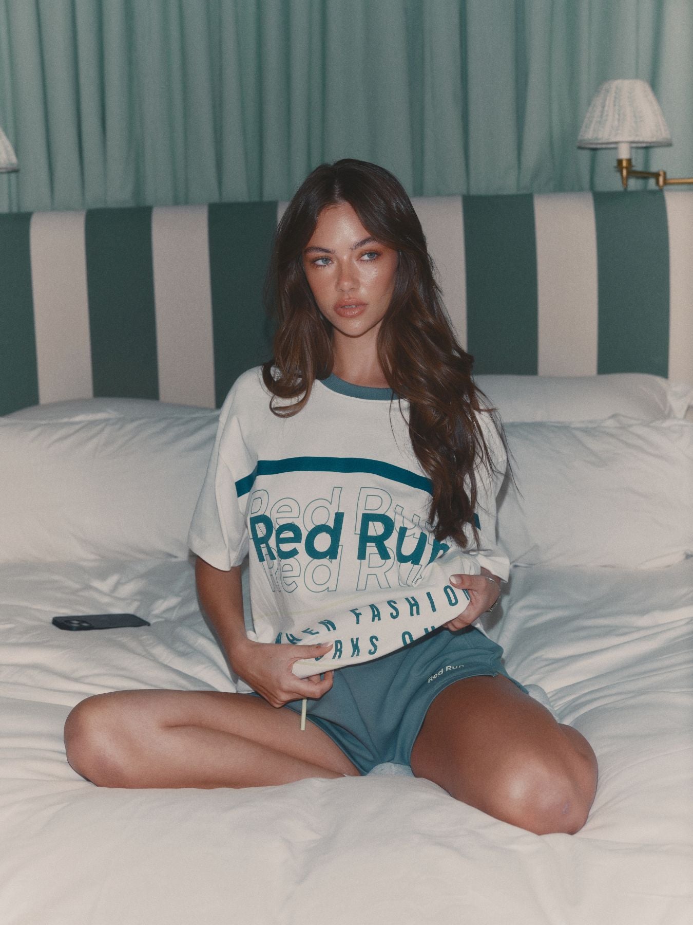 Woman sitting on a bed wearing a 'Red Rum' t-shirt and shorts.